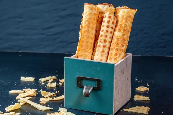 Waffelrollen in türkisfarbener Box auf blauem Tisch, umgeben von Krümeln