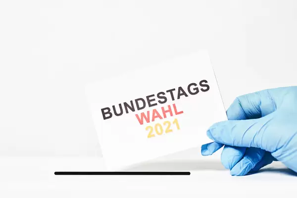 Wahl und Corona: Hand mit Einmalhandschuh wirft Zettel mit Schrift "Bundestagswahl 2021" in die Wahlurne ein