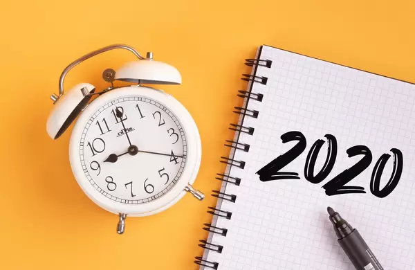 Wecker und ein Heft mit ‘2020’ Text mit einem Filzstift drauf vor gelbem Hintergrund