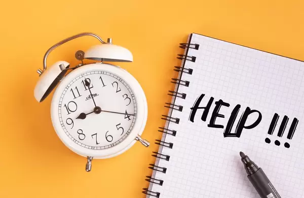 Wecker und ein Heft mit ‘Help!!!’ Text mit einem Filzstift drauf vor gelbem Hintergrund