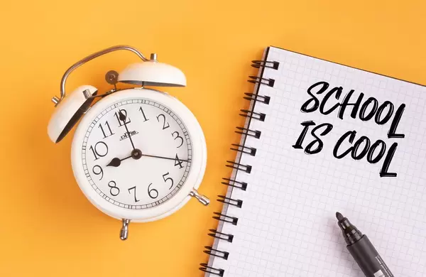 Wecker und ein Heft mit ‘School is cool’ Text mit einem Filzstift drauf vor gelbem Hintergrund