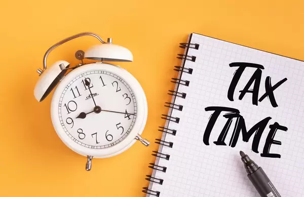 Wecker und ein Heft mit ‘Tax time’ Text mit einem Filzstift drauf vor gelbem Hintergrund