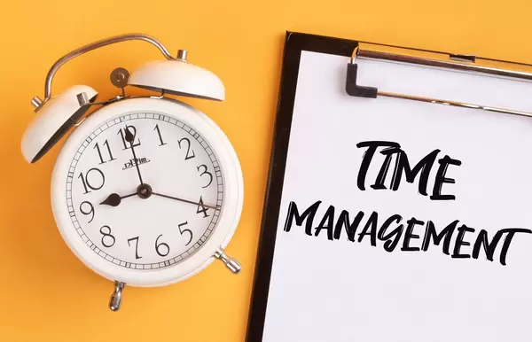 Wecker und ein Klemmbrett mit dem handgeschriebenen Text "Time Management / Zeitmanagement", vor gelbem Hintergrund