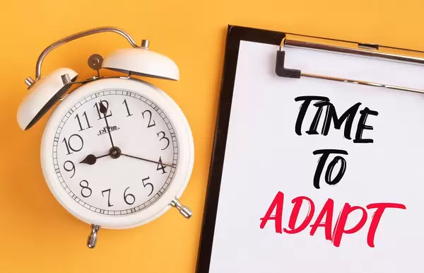 Wecker und ein Klemmbrett mit dem handgeschriebenen Text "Time to adapt / Zeit für Anpassung", vor gelbem Hintergrund