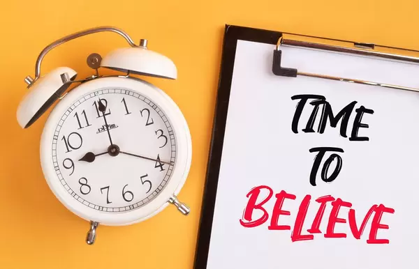 Wecker und ein Klemmbrett mit dem handgeschriebenen Text "Time to beliebe / Zeit zu glauben", vor gelbem Hintergrund