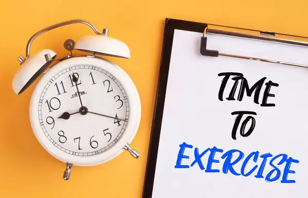 Wecker und ein Klemmbrett mit dem handgeschriebenen Text "Time to exercise / Zeit zum Trainieren", vor gelbem Hintergrund