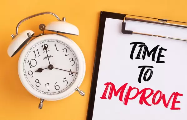 Wecker und ein Klemmbrett mit dem handgeschriebenen Text "Time to improve / Zeit sich zu verbessern", vor gelbem Hintergrund