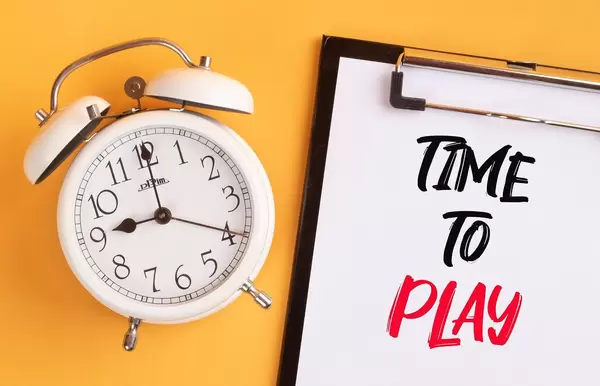 Wecker und ein Klemmbrett mit dem handgeschriebenen Text "time to play / Zeit zum Spielen", vor gelbem Hintergrund