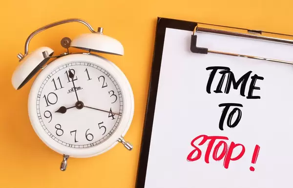 Wecker und ein Klemmbrett mit dem handgeschriebenen Text "Time to stop / Es ist Zeit aufzuhören", vor gelbem Hintergrund