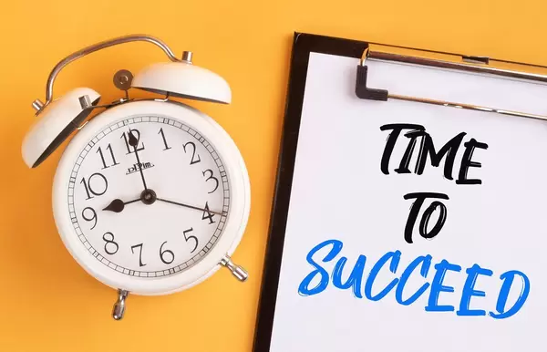 Wecker und ein Klemmbrett mit dem handgeschriebenen Text "Time to succeed / Zeit für Erfolg", vor gelbem Hintergrund