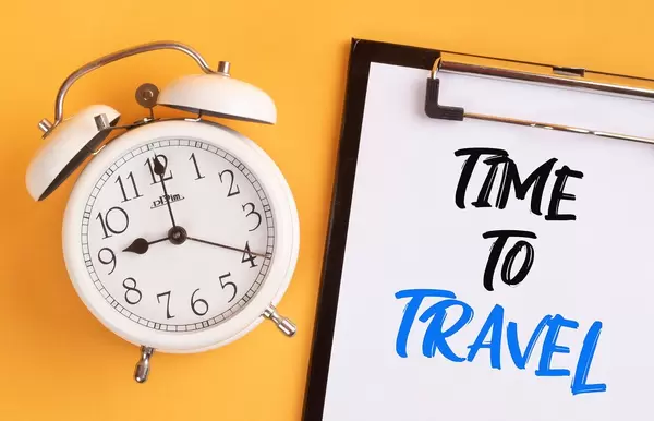Wecker und ein Klemmbrett mit dem handgeschriebenen Text "Time to travel / Zeit zum Reisen", vor gelbem Hintergrund