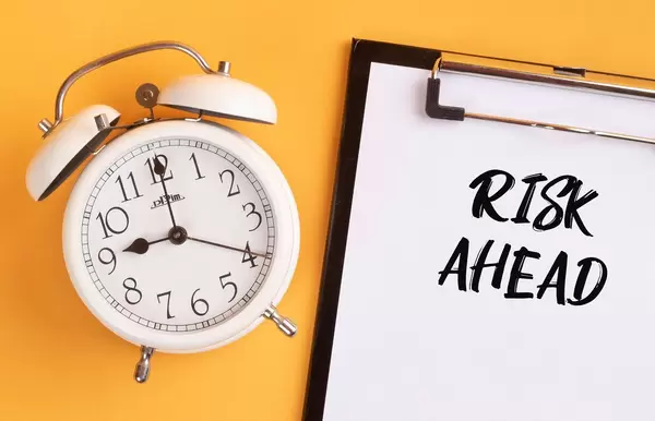 Wecker und ein Klemmbrett mit dem Text "Risk ahead / auf einen zukommendes Risiko", vor gelbem Hintergrund
