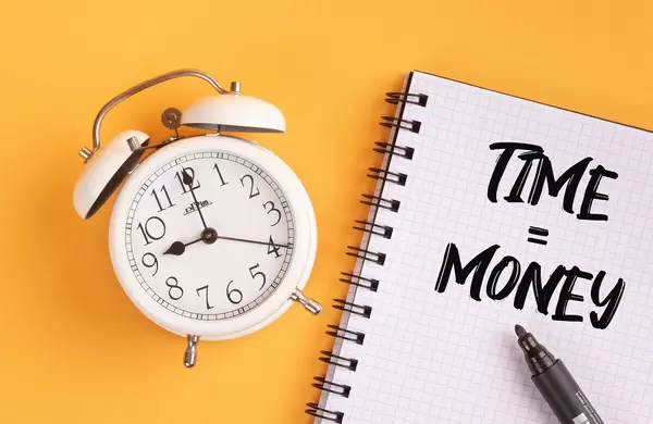 Wecker und ein Notizblock mit dem Text "Time is money / Zeit ist Geld", mit einem Filzstift vor gelbem Hintergrund