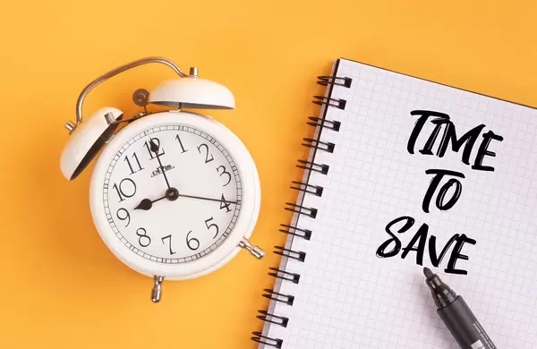 Wecker und ein Notizblock mit dem Text "time to Save / Zeit zu sparen", mit einem Filzstift vor gelbem Hintergrund