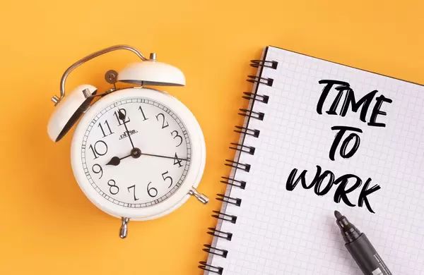 Wecker und ein Notizblock mit dem Text "time to work / Zeit zu arbeiten", mit einem Filzstift vor gelbem Hintergrund