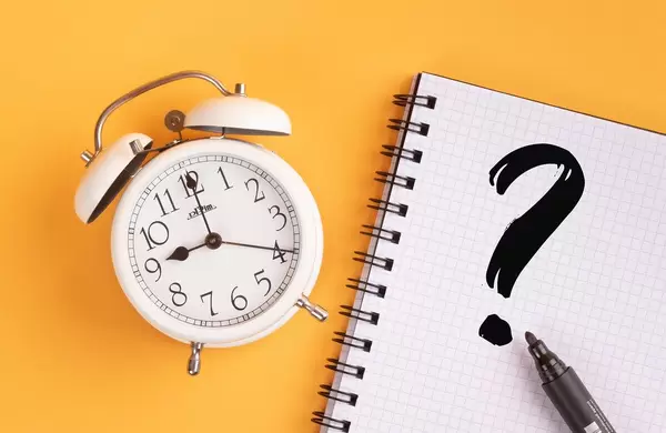 Wecker und ein Notizblock mit einem Fragezeichen und einem schwarzen Filzstift vor gelbem Hintergrund