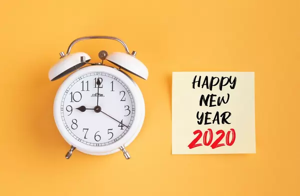 Wecker und ein Zettel mit ‘Happy new year 2020’ Text vor gelbem Hintergrund
