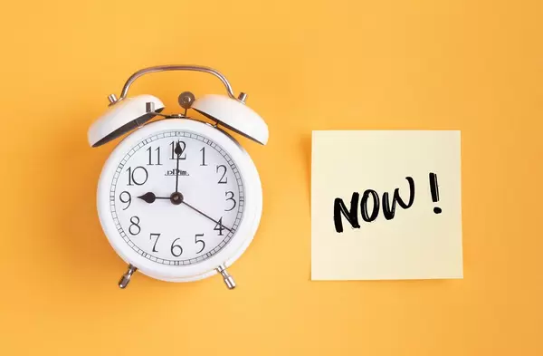 Wecker und ein Zettel mit ‘Now!’ Text vor gelbem Hintergrund