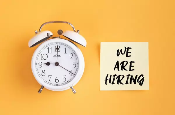Wecker und ein Zettel mit ‘We are hiring’ Text vor gelbem Hintergrund