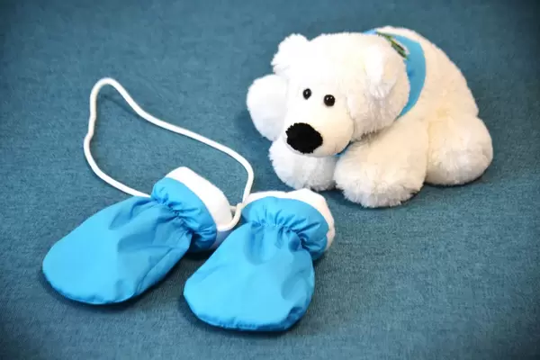 Weißer Plüsch-Eisbär mit blauem Dreieckstuch vor hellblau-weißen Baby-Fäustlingen mit Halskordel auf blauem Teppich