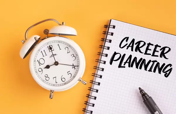 Weißer Wecker und ein Heft mit ‘Career planning’ Text mit einem Filzstift drauf vor gelbem Hintergrund