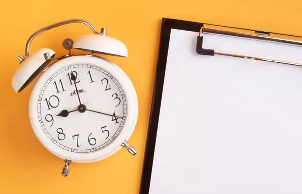 White alarm clock on yellow background with empty clipboard
