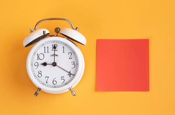 White alarm clock on yellow background with empty red sticky note