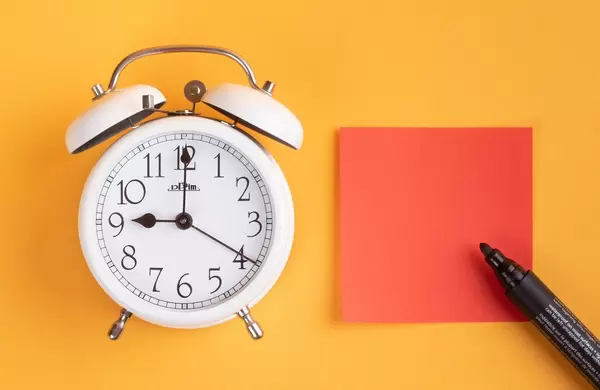 White alarm clock on yellow background with empty sticky note and pen