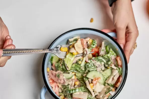 Woman's hand holds a fork with mixed salad with sweetcorn, cucumber, lettuce, mushrooms from a pot