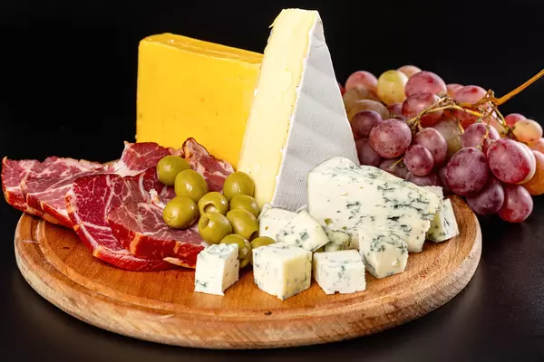 Wooden kitchen board with sliced delicatessen cheeses, ham, olives and grapes