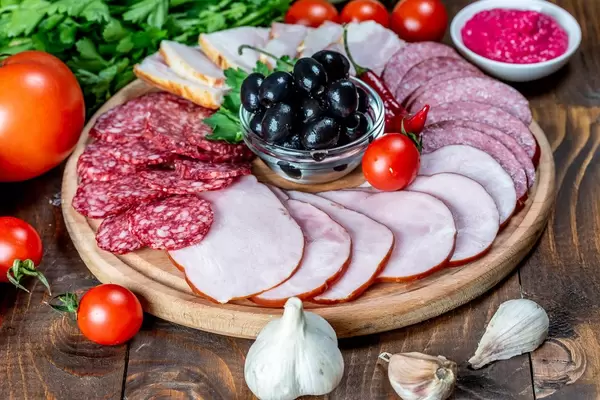 Wurstteller mit frischer Salami und geräuchertem Schinken, präsentiert zwischen frischem Gemüse und Knoblauch auf einem Holztisch