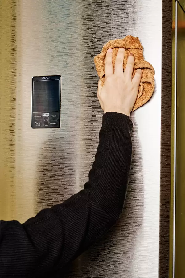 Young woman cleaning stainless steel refrigerator with cloth