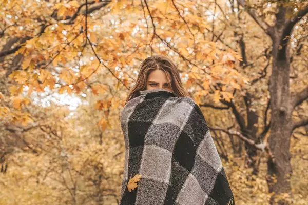Young woman walking in autumnal park, hiding her face with warm woolen plaid