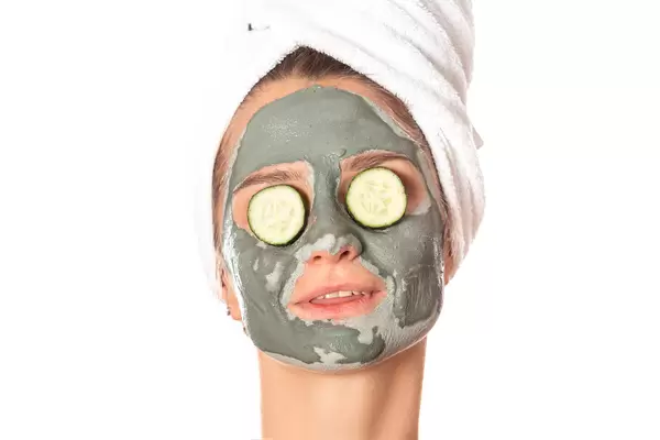 Young woman with a clay mask, covering her eyes with cucumbers on white background