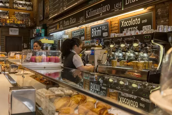Zwei Angestellte in Arbeitskleidung, hinter der Ladentheke einer spanischen Bäckerei in Barcelona mit allerlei Gebäck und Kaffee