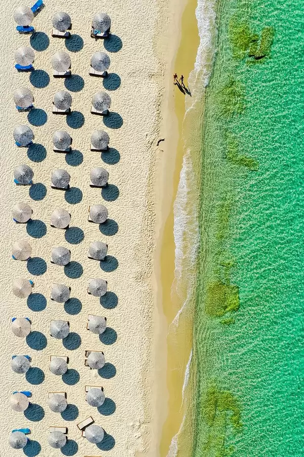 Zwei Menschen laufen den Sandstrand entlang. Drohnenaufnahme mit Strandschirmen auf Naxos