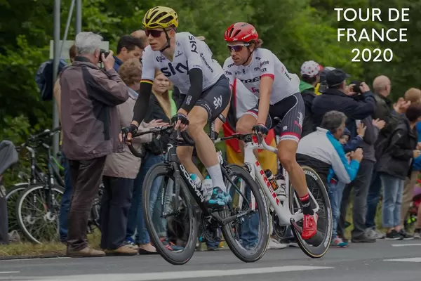 Zwei Radrennfahrer auf ihren Rennrädern, mit Zuschauern am Straßenrand, neben der Aufschrift "Tour de France 2020"