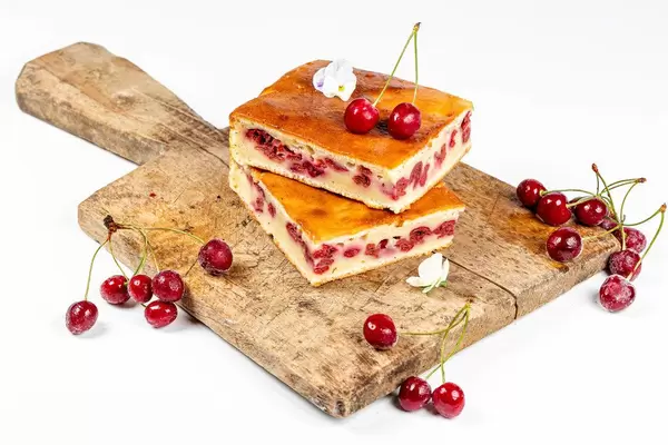 Zwei Stück Kirschkuchen auf einem hölzernen Schneidebrett mit frischen Kirschen und Blumen vor weißem Hintergrund