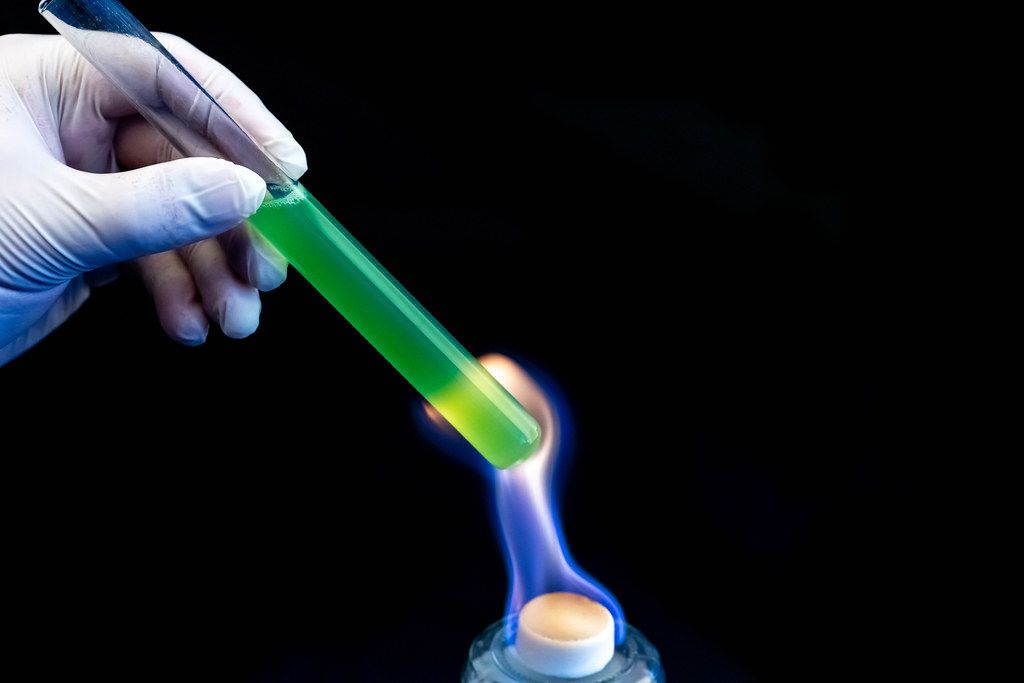 A laboratory test tube with a green solution in a man's hand is heated over a fire