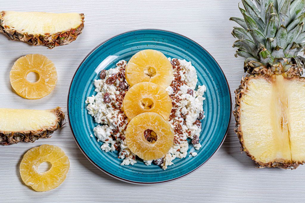 A plate of cottage cheese with flax seeds and dried fruits on a white wooden background with pieces of fresh pineapple