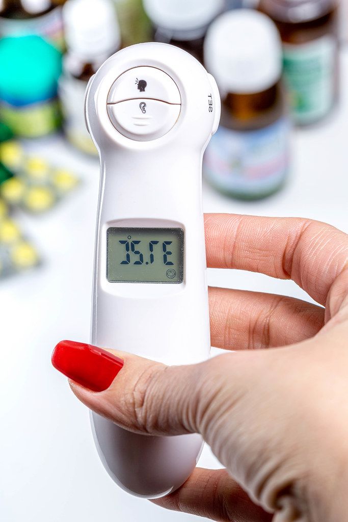 A woman's hand holds an electric thermometer with a temperature of 37.2, behind the medical supplies background (Flip 2019)