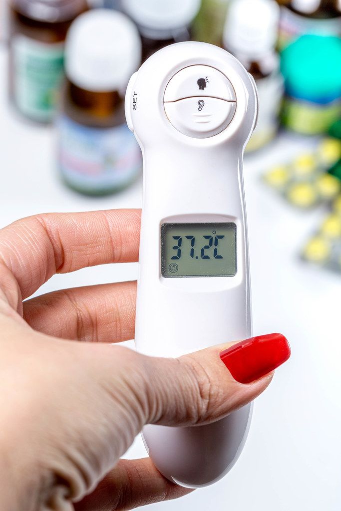 A woman's hand holds an electric thermometer with a temperature of 37.2, behind the medical supplies background