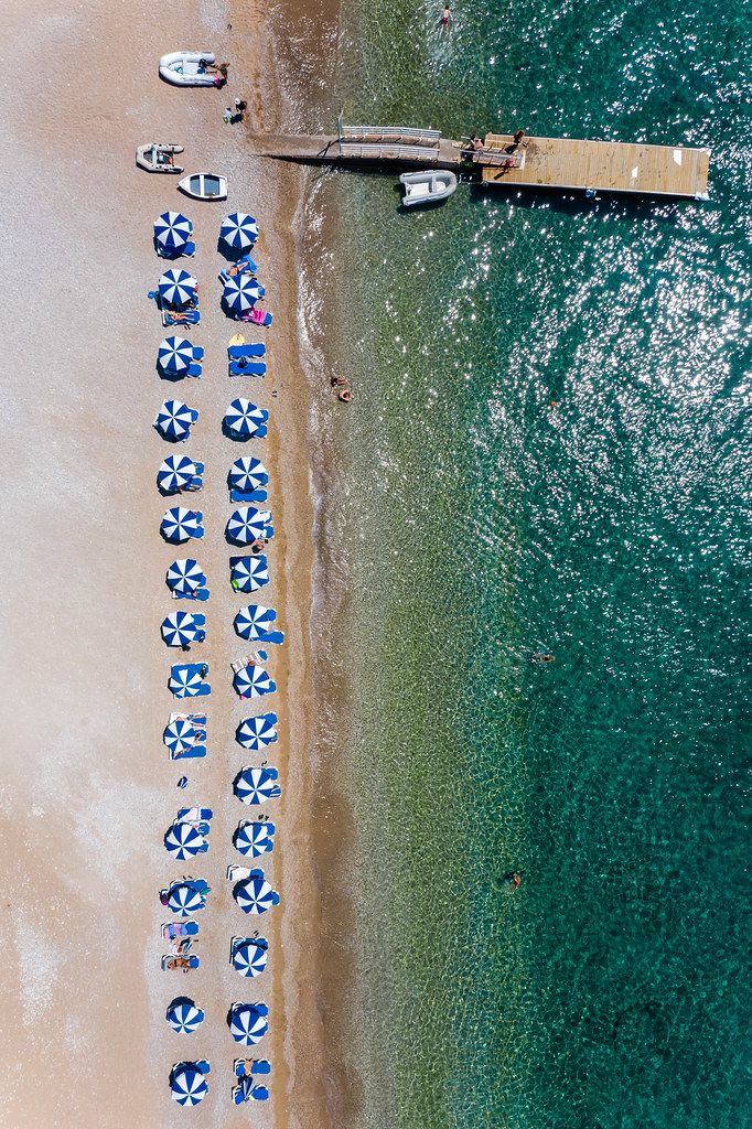 Aerial view shows neatly placed parasols in blue and white, in two rows, at the sandy beach of Agii Anargiri & green sea