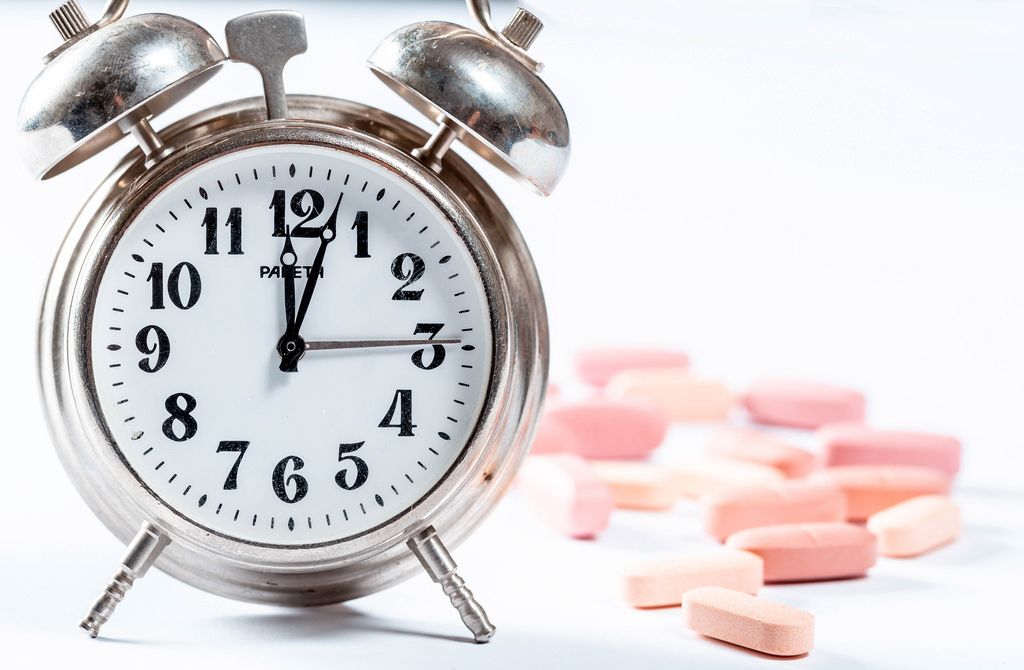 Alter Wecker aus Metall neben rosaroten Tabletten mit Schärfentiefe vor weißem Hintergrund