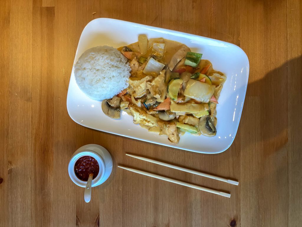 Asiatisches Gericht mit Hühnchen, Pilzen, Karotten und weiterem Gemüse in Currysoße, dazu Reishügel auf Teller, Chilipaste in weißem Gefäß und Essstäbchen auf Holztisch