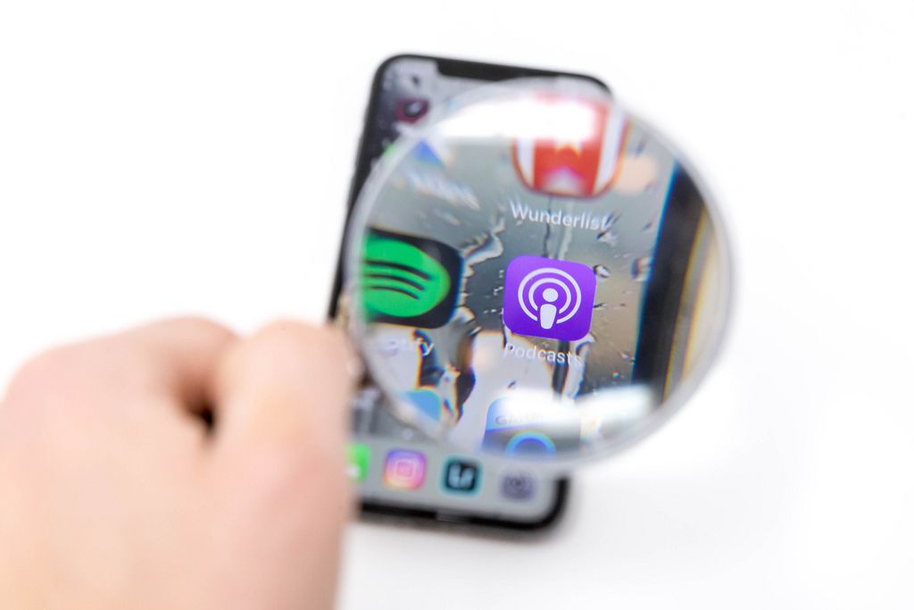 Auf Mobiltelefon durch Lupe betrachtetes Podcast App Logo neben Spotify Icon vor weißem Hintergrund