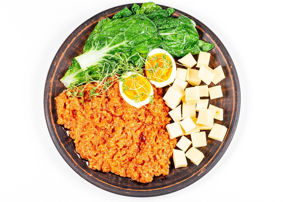 Aufnahme von oben: Gemüse mit gekochtem Ei, Käsewürfel und Zucchinikaviar auf einem braunen Teller vor weißem Hintergrund