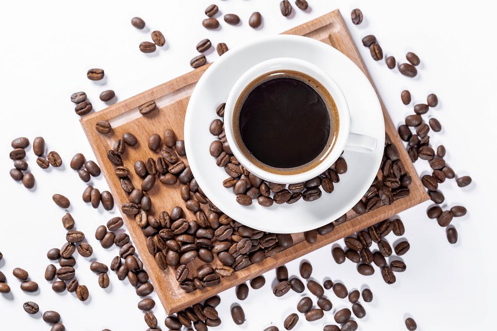 Aufsicht auf eine Tasse Kaffee mit Kaffeebohnen auf einem Holzbrettchen auf weißem Hintergrund