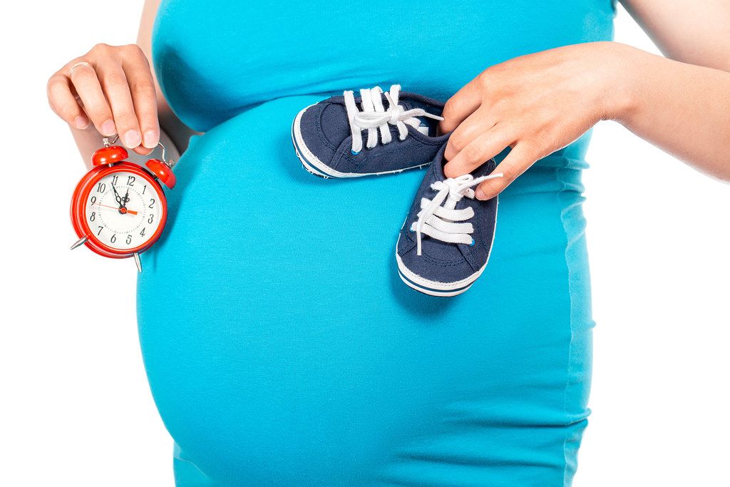 Beautiful small blue shoes at the pregnant woman's belly and red alarm in hand