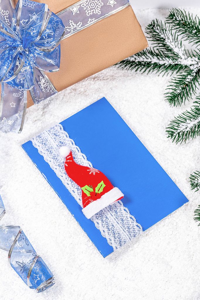 Blaue Weihnachtsgrußkarte mit roter Mütze und Tannenzweigen vor einem Geschenk im Schnee